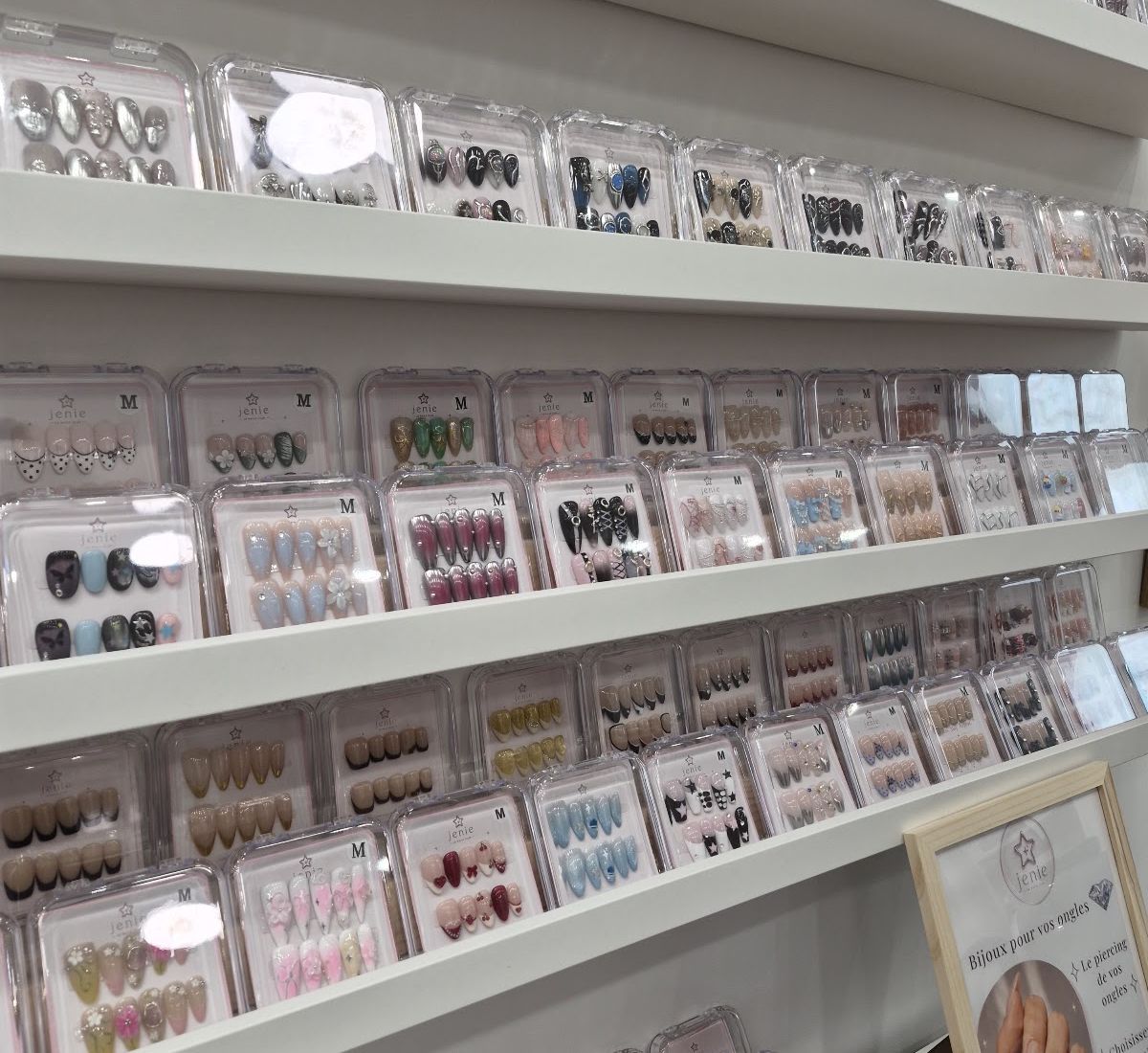 Le mur de boîtiers transparents de Jenie : des rangées de press-on sur étagères blanches, en perspective.